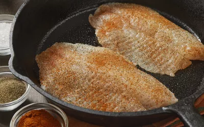 Two seasoned fish fillets being seared in a cast iron skillet with spices nearby.
