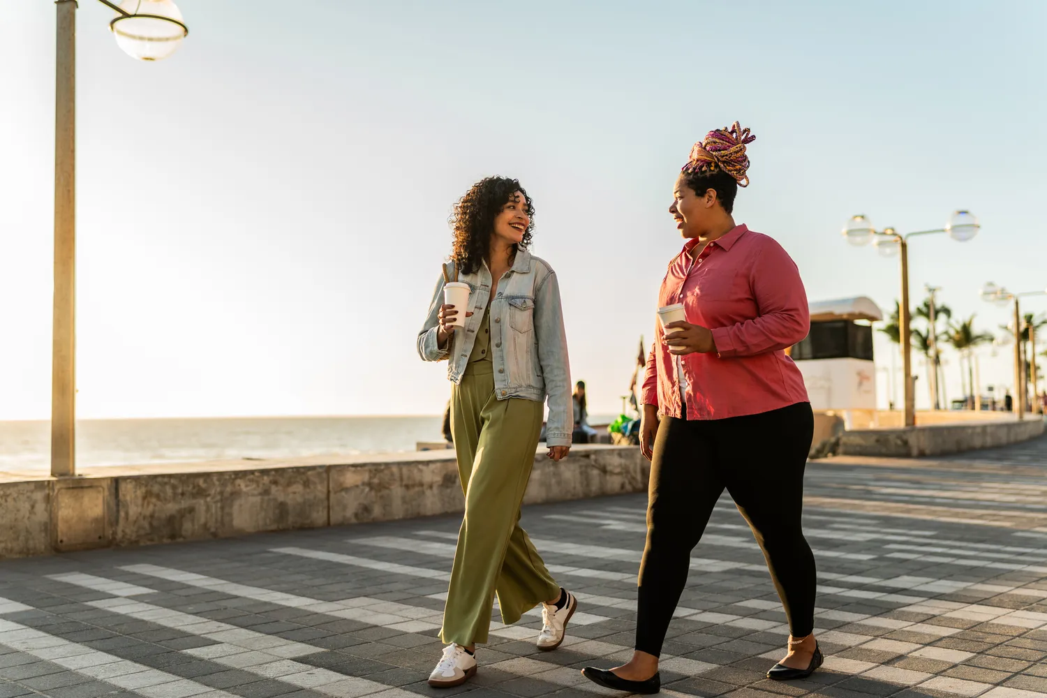 Two women laughing and walking together on a sunny seaside boardwalk with coffee.