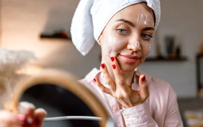 A woman in a towel applying facial cream while looking in a bathroom mirror.