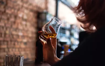 A person holding a glass of amber liquor in a bar setting.