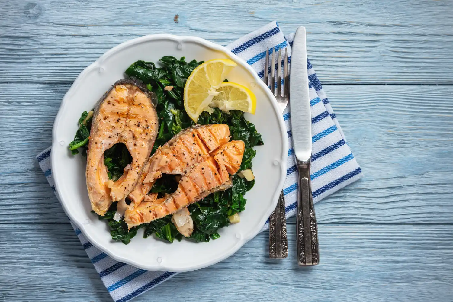 Two grilled salmon fillets served with fresh spinach and lemon on a blue wooden surface.