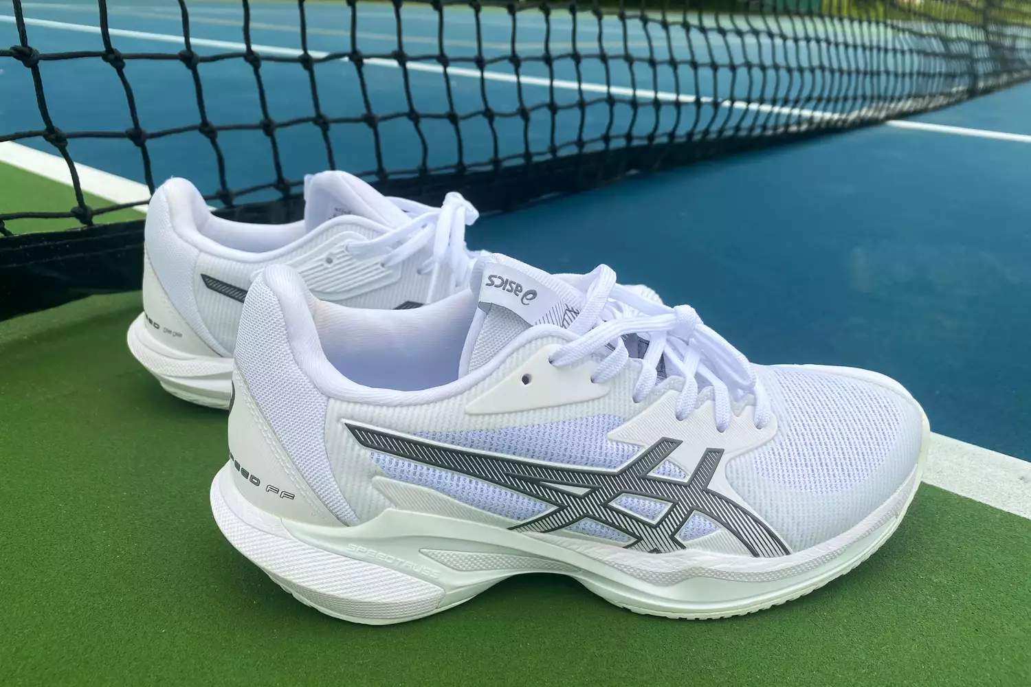 White Asics Solution Speed FF 3 shoes resting on a green tennis court near the net.
